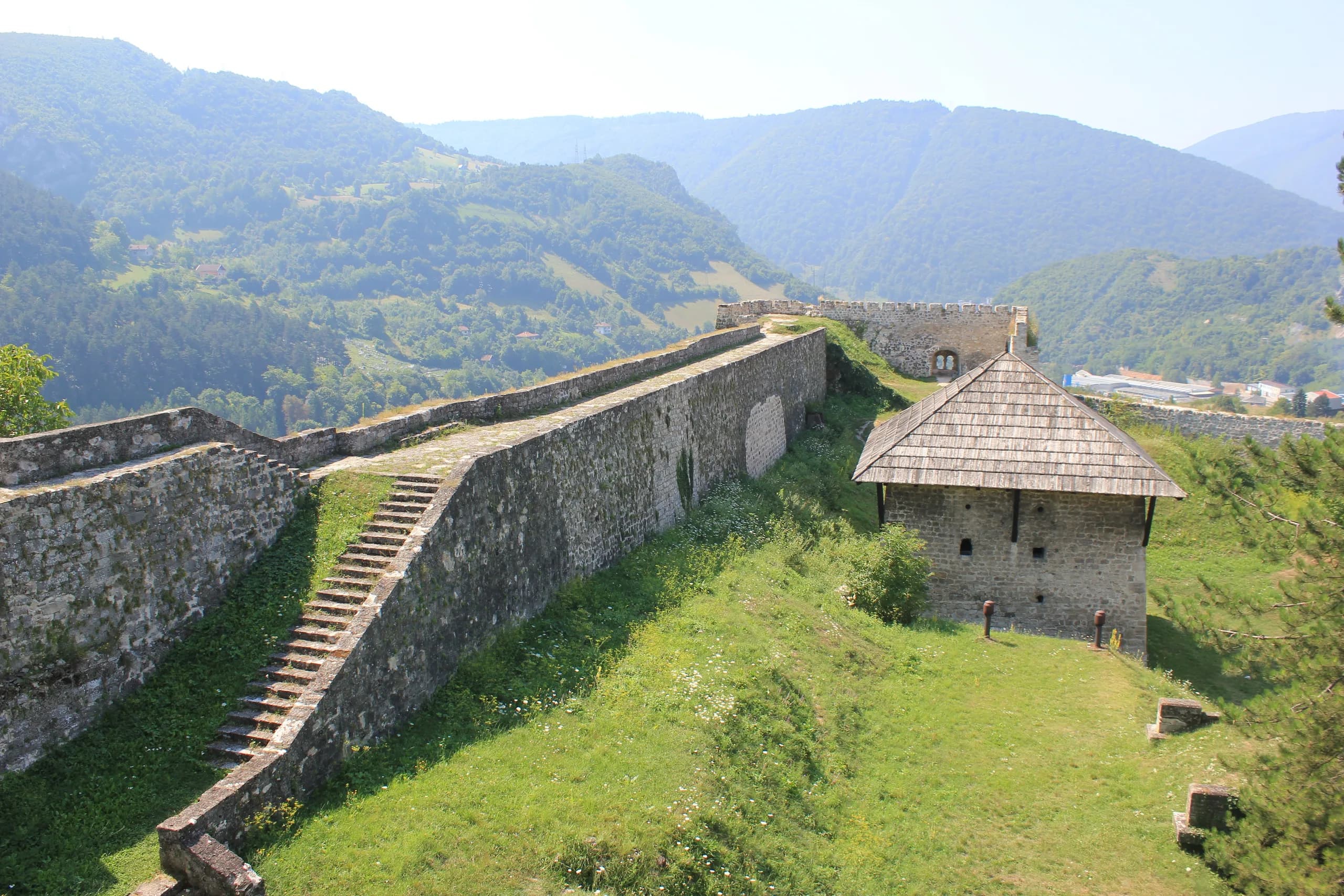 Mauern und Treppe der Festung Jajce, daneben ein Steinbau mit Schindeldach, dahinter bewaldete Berge