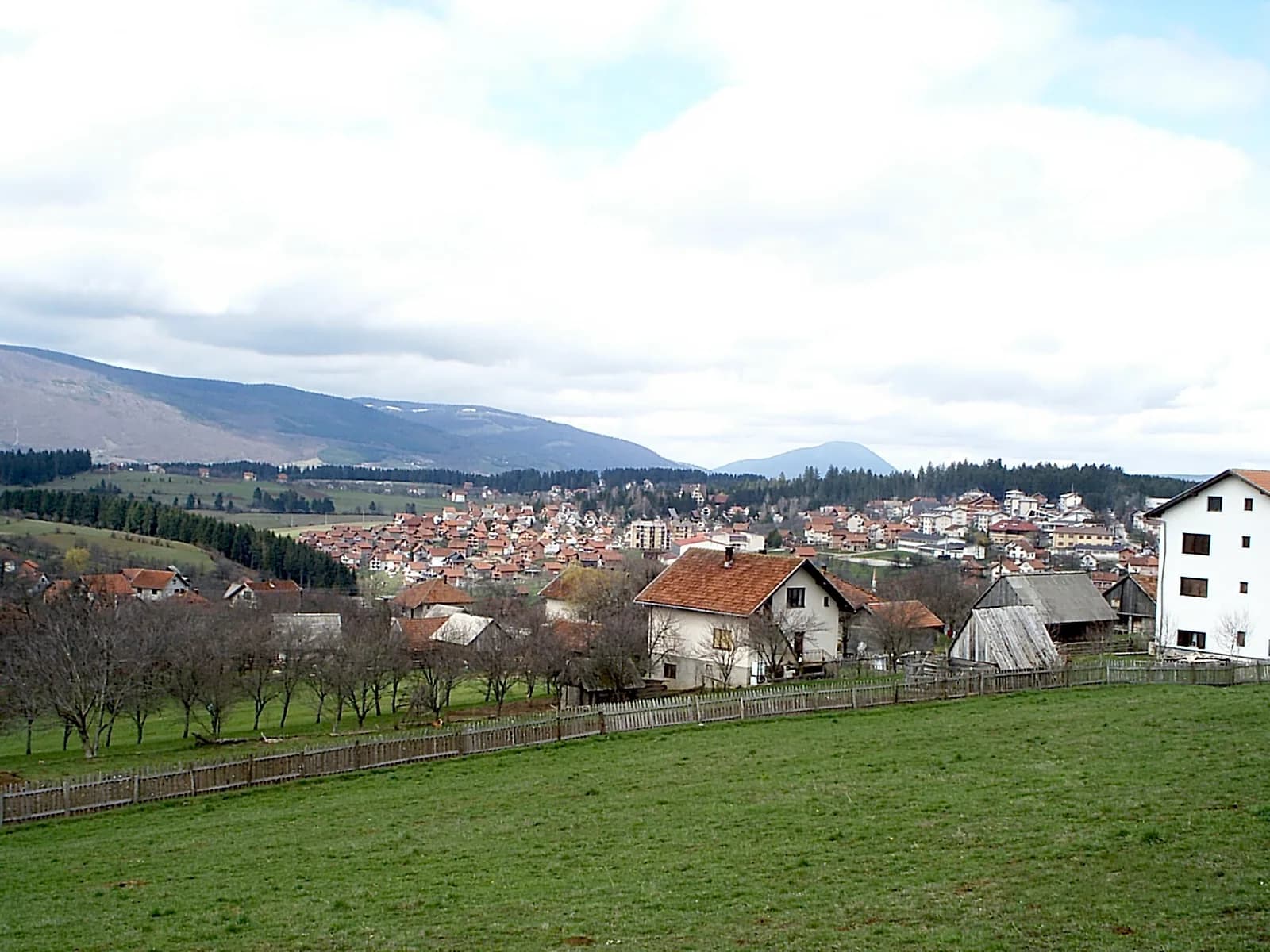 Blick über eine Wiese und kahle Bäume auf die Häuser von Kneževo, dahinter Berge