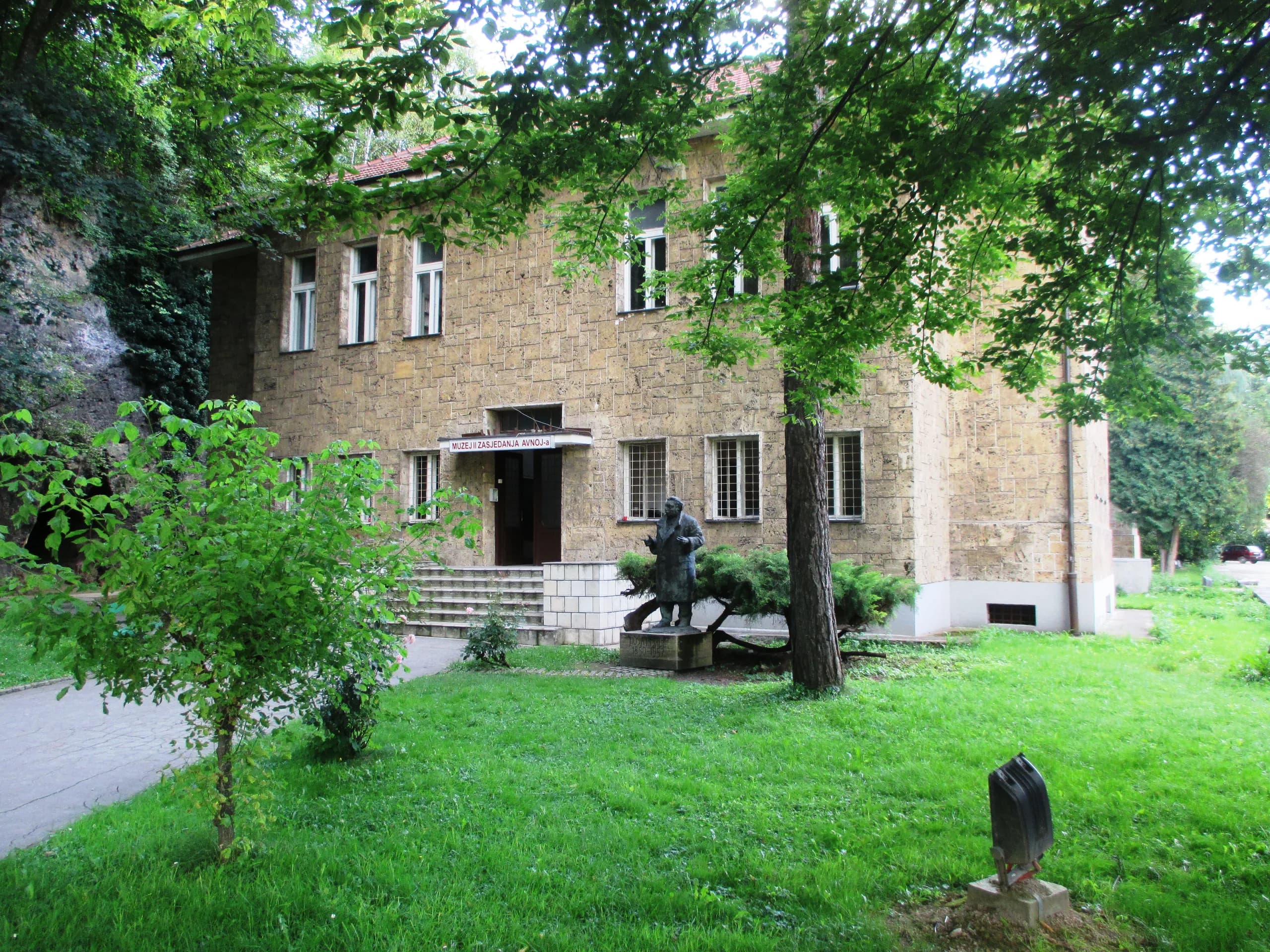 Das Steingebäude des AVNOJ-Museums in Jajce hinter Bäumen und Rasen, vor dem Eingang eine Statue